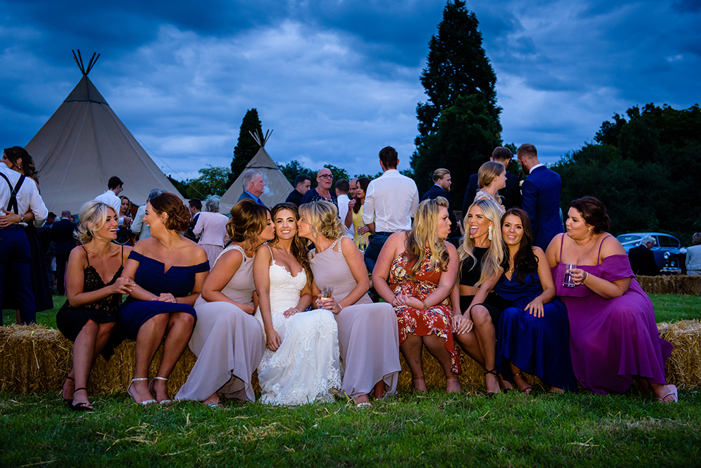 bride and friends sit on hay bale at hertfordshire tipi wedding